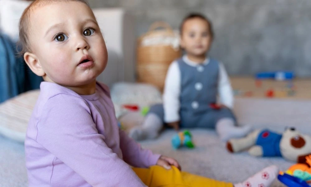 Deux jeunes enfants jouent assis sur le sol d’une pièce, entourés de jouets.