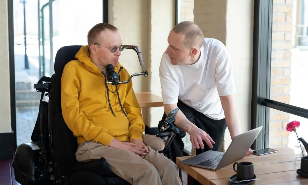 Deux personnes, l'une en fauteuil roulant et l'autre debout, travaillent ensemble sur un ordinateur portable dans un bureau lumineux.