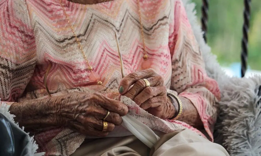Une personne âgée portant des bijoux aux mains et au cou, essuie ses lunettes dans son pull.