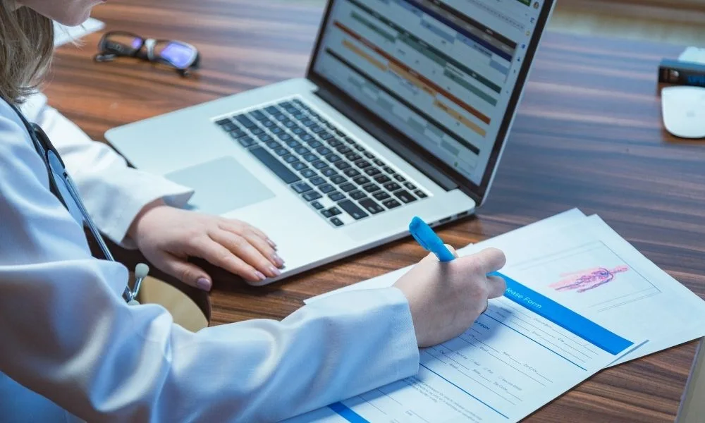 Une femme médecin est assise à un bureau, travaillant sur des documents tout en regardant un ordinateur portable ouvert devant elle.