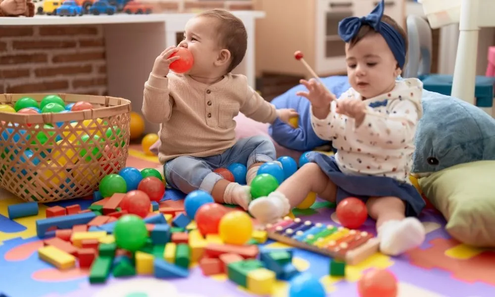 Deux bébés jouent joyeusement avec des jouets colorés sur un tapis, entourés de blocs et de boules.