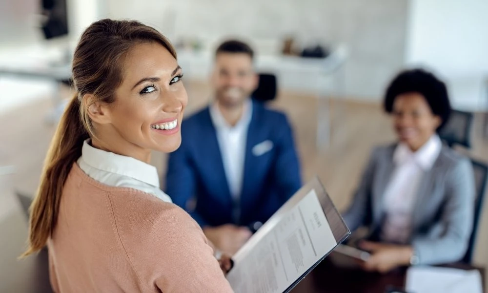 Une femme souriante tient un document en main, entourée de collègues dans une ambiance professionnelle.