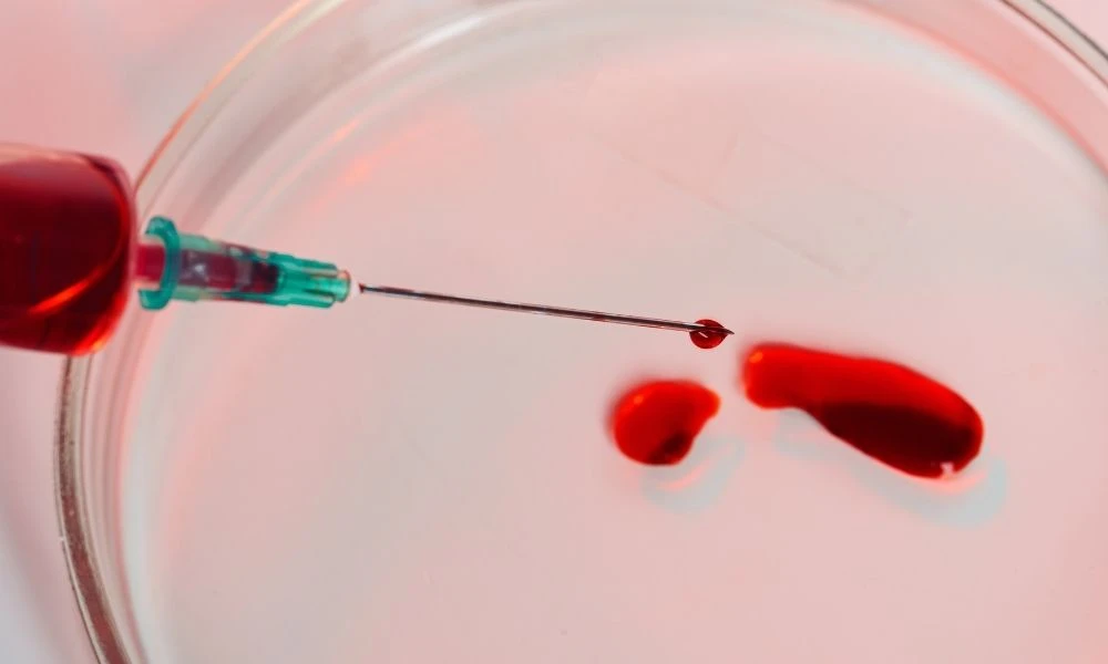 Une seringue injecte une goutte de sang dans une coupelle en verre, probablement pour un test ou une analyse.