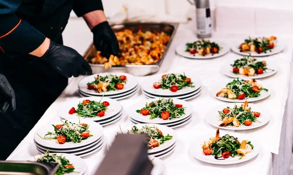 Un chef en tenue professionnelle prépare des assiettes de salades garnies avec soin sur une table blanche.