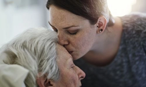 Une jeune femme embrasse tendrement le front d'une personne âgée allongée, exprimant affection et réconfort.