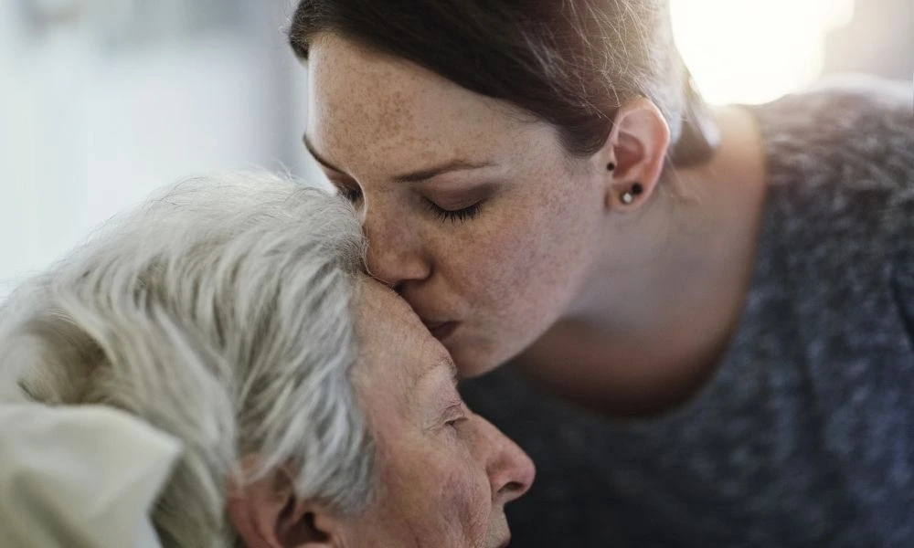 Une jeune femme embrasse tendrement le front d'une personne âgée allongée, exprimant affection et réconfort.