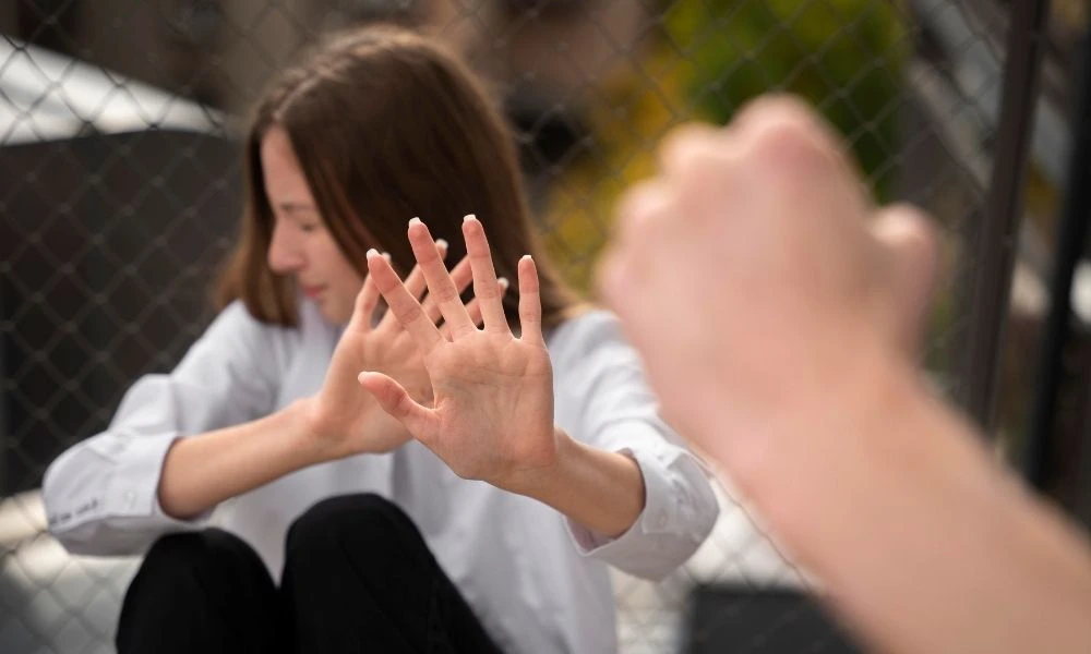 Une personne assise, lève sa main droite avec les doigts écartés devant son visage pour se protéger, une autre personne s’avance le poing levé devant elle.