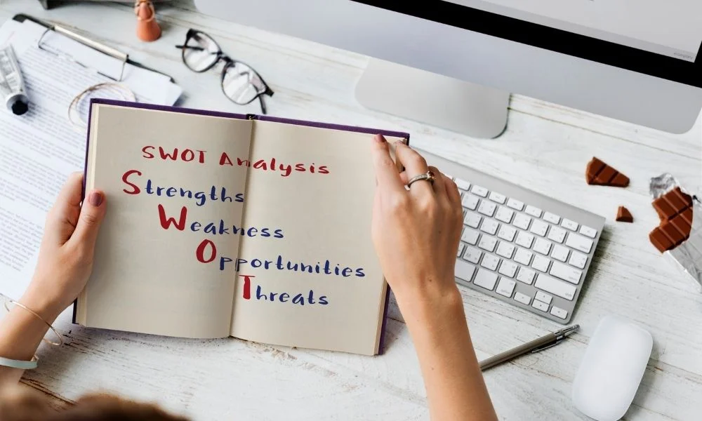Une personne tient un carnet ouvert sur une table de travail, où sont inscrits les mots "SWOT Analysis", "Strengths", "Weakness", "Opportunities" et "Threats", dans un environnement professionnel avec un ordinateur, des lunettes et des objets décoratifs.