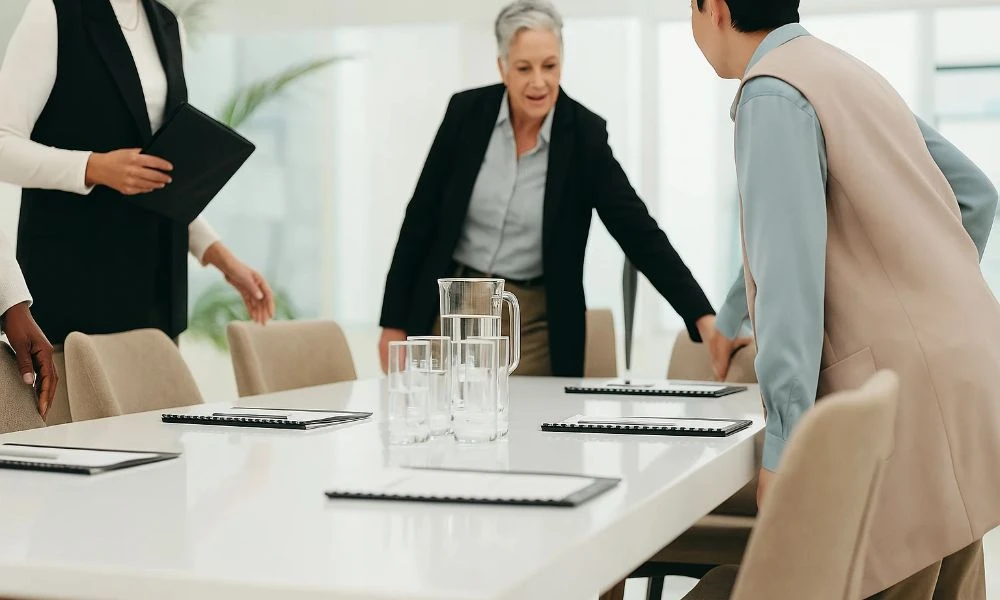 Des personnes en tenue professionnelle s'installent pour une réunion qui va se dérouler autour d'une table sur laquelle est disposée des documents et des verres d'eau.