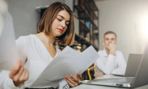 Une femme en chemise blanche examine des documents dans un bureau, tandis qu'un homme assis derrière elle semble réfléchir.