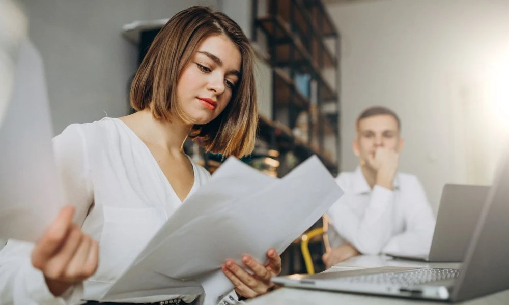 Une femme en chemise blanche examine des documents dans un bureau, tandis qu'un homme assis derrière elle semble réfléchir.
