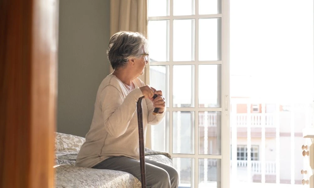 Une résidente d’EHPAD assise sur son lit, regardant pensivement par la fenêtre, canne en main.