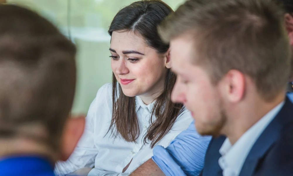 Cette image montre une femme souriante en chemise blanche travaillant en équipe avec des collègues dans un cadre professionnel.