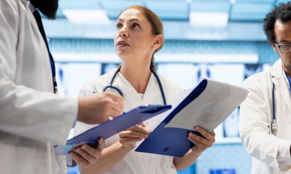 L'image montre une équipe de professionnels de santé discutant et prenant des notes sur des dossiers dans un environnement hospitalier.