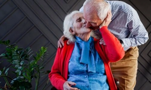 Couple de personnes âgées s'embrasse tendrement. Madame est assise et monsieur se penche pour l'embrasser.