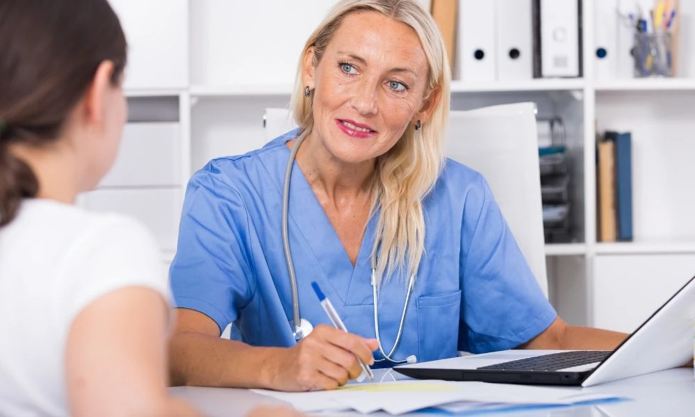 L'image montre une professionnelle de santé en blouse bleue discutant avec une personne.