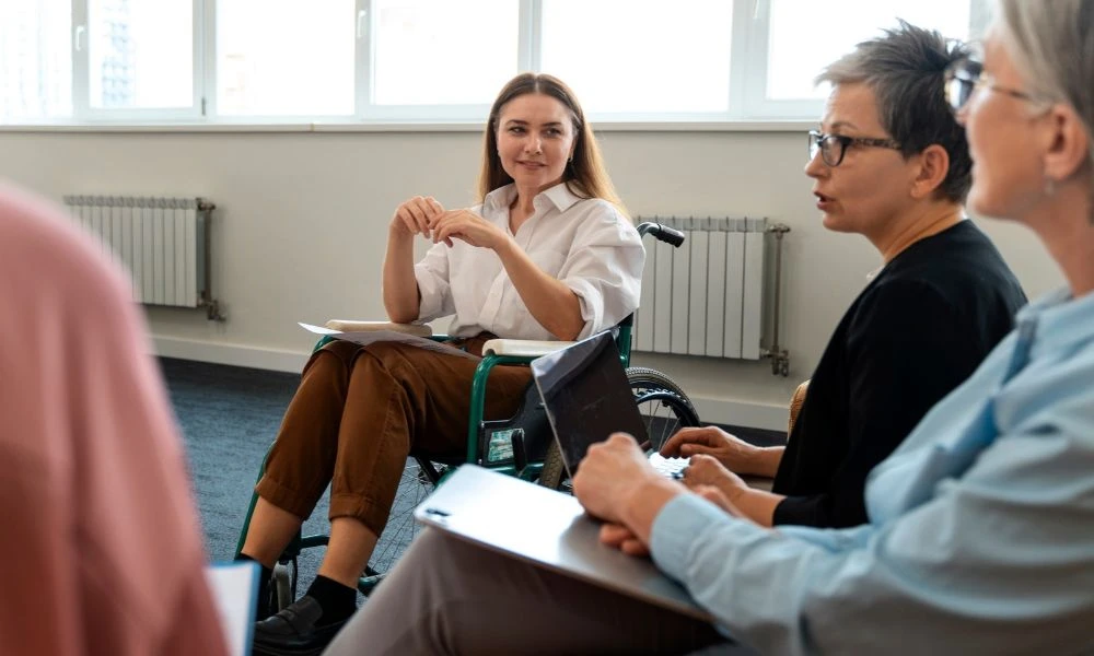 L'image montre un groupe de femmes en discussion dans une salle lumineuse, dont l'une est en fauteuil roulant et semble diriger l'échange.