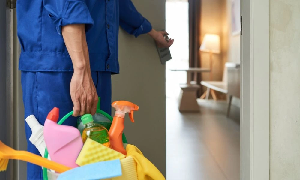 Cette image montre une personne chargée de l’entretien tenant un seau rempli de produits de nettoyage et d'accessoires. Parmi ces objets, on distingue des pulvérisateurs, des éponges et des bouteilles de détergent.