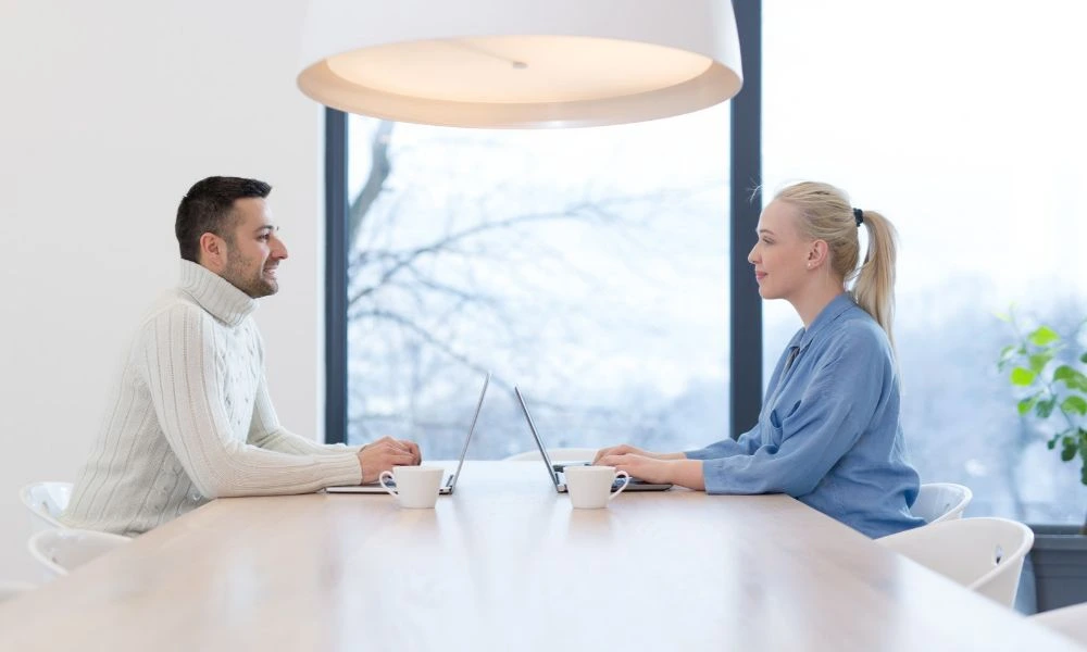 Cette image montre deux personnes assises face à face de chaque côté d'une table. Les deux individus sont engagés dans une conversation professionnelle.