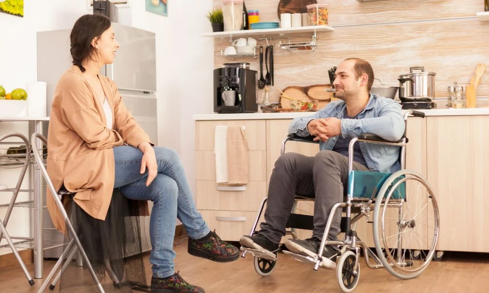 Cette image montre un homme en fauteuil roulant discutant avec une femme assise sur une chaise pliante dans une cuisine.