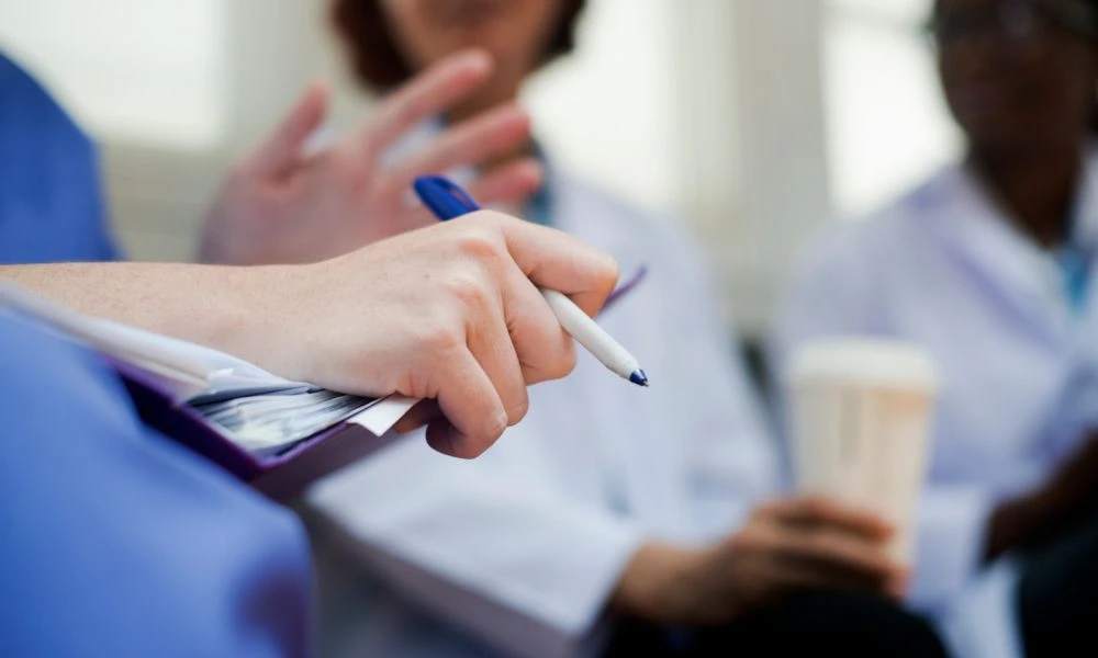 Cette image montre une scène de discussion en milieu médical. Au premier plan, une main tient un stylo bleu et semble prête à écrire sur un carnet ou une tablette de notes.