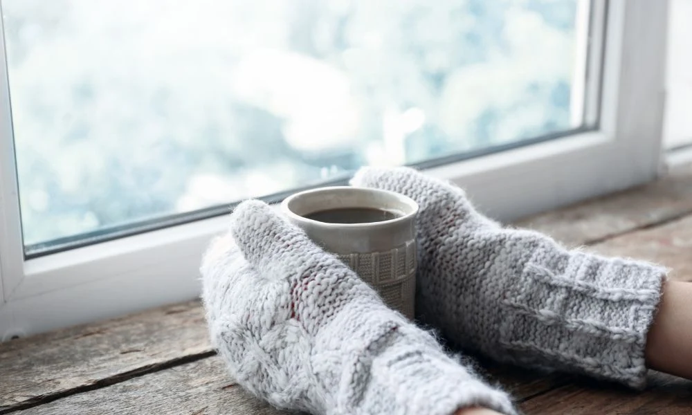 L'image montre des mains couvertes de gants gris en tricot épais, tenant une tasse de boisson chaude près d'une fenêtre.