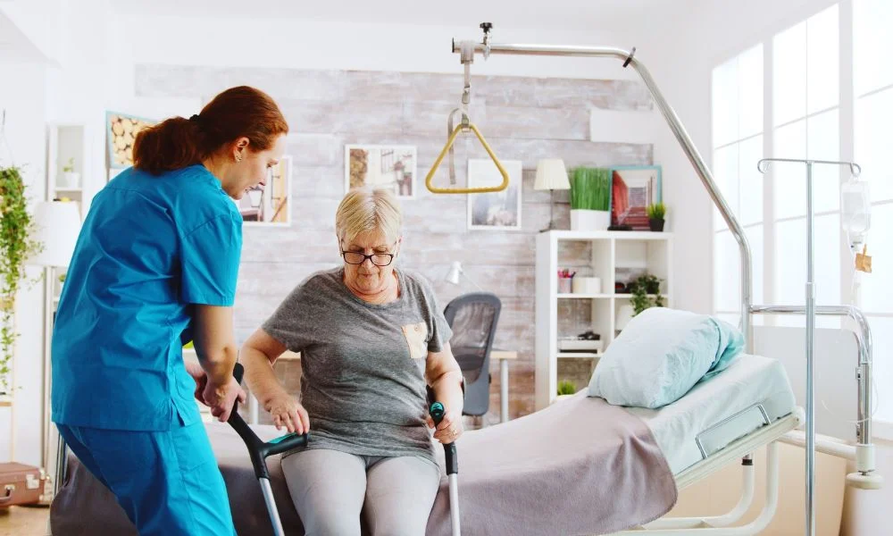Sur cette image, on voit une infirmière aider une patiente âgée à se lever de son lit médicalisé en soins à domicile.