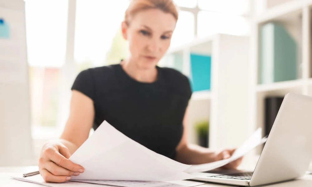 Une femme en tenue professionnelle consulte des documents papier à son bureau, avec un ordinateur portable ouvert à côté d'elle.