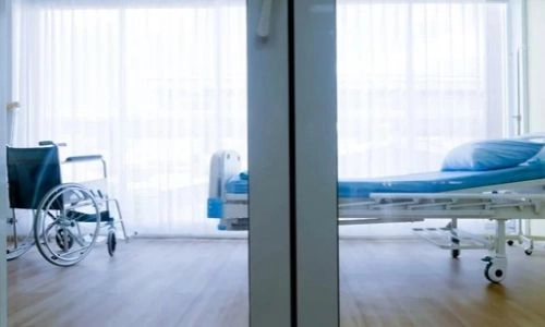 L'image montre une chambre d'hôpital lumineuse avec un grand rideau blanc qui laisse entrer la lumière naturelle. Au centre, on peut voir un lit médicalisé avec une couverture et, à gauche, un fauteuil roulant à côté du lit.