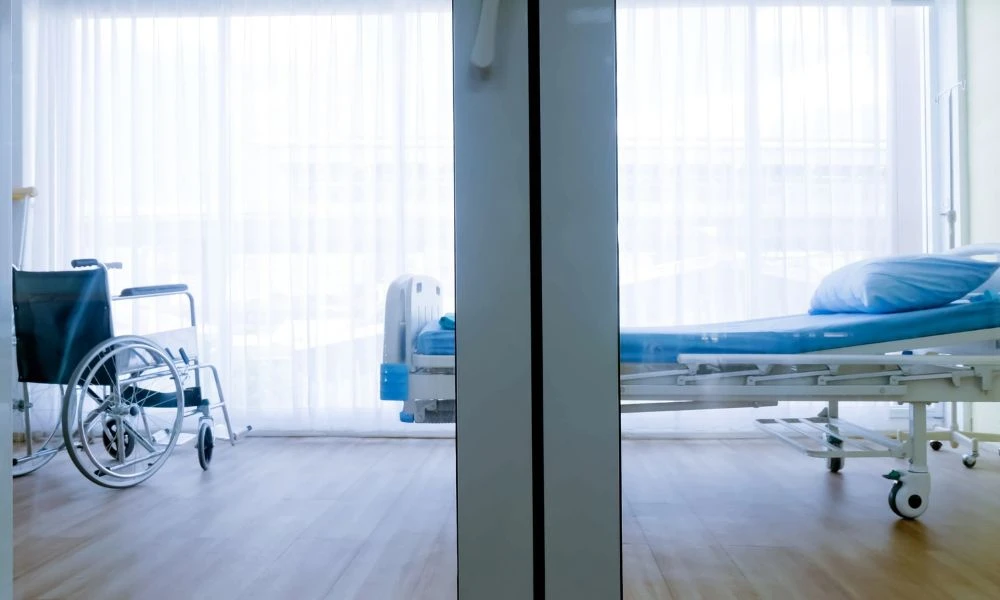 L'image montre une chambre d'hôpital lumineuse avec un grand rideau blanc qui laisse entrer la lumière naturelle. Au centre, on peut voir un lit médicalisé avec une couverture et, à gauche, un fauteuil roulant à côté du lit.