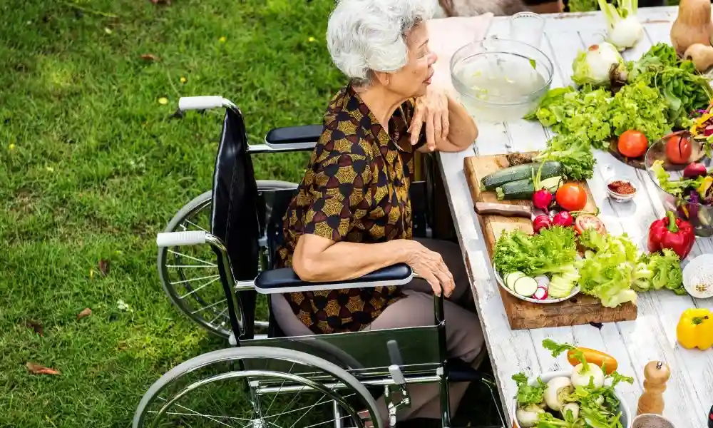 Personne âgée assise dans un fauteuil roulant à l'extérieur, près d'une table remplie de légumes et de légumes verts frais.