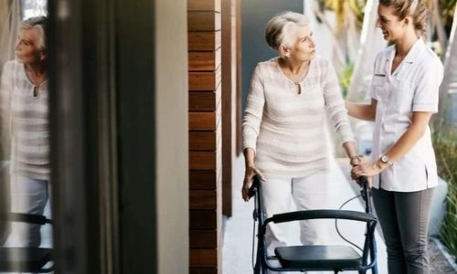 Une femme âgée utilise un déambulateur et elle est aidée par une soignante ou une aide-soignante.
