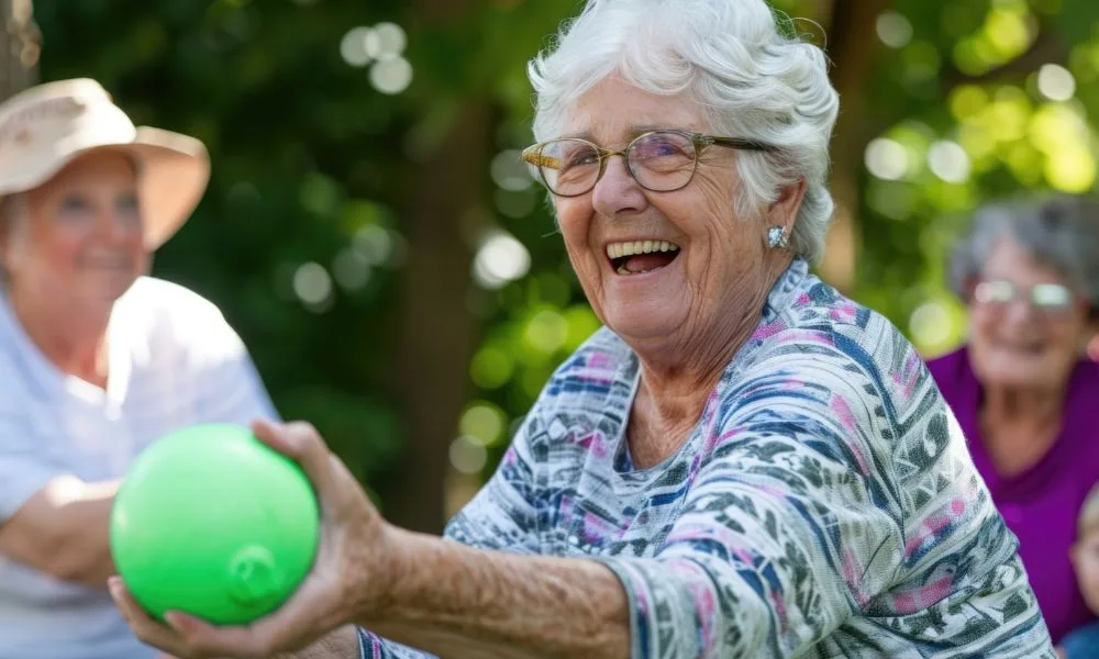 Scène joyeuse en plein air où plusieurs personnes âgées participent à une activité ludique. Au premier plan, une femme âgée souriante tient une balle verte dans la main.
