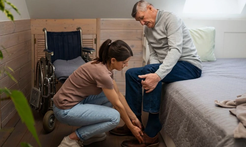 Femme jeune en train d'aider un homme âgé à s'habiller. L'homme est assis sur un lit, tandis que la femme est accroupie devant lui, en train de lui mettre des chaussures. Un fauteuil roulant est visible en arrière-plan, suggérant que l'homme a des difficultés à se déplacer.