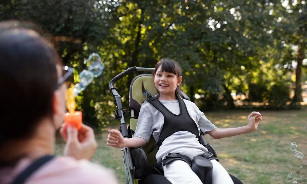 Jeune fille en situation de handicap fauteuil roulant souriante. Elle partage un moment de joie dans un parc avec une personne qui lui envoie des bulles de savon.