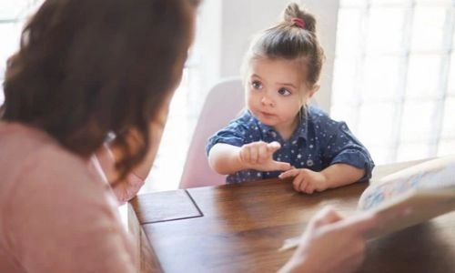 Une petite fille assise à une table, pointe le doigt en s'exprimant face à une adulte qui l'écoute attentivement, dans une pièce lumineuse.