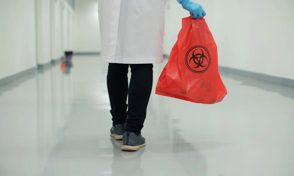 Professionnel de soin muni de gants bleus marche dans un couloir d'hôpital. Il porte à la main droite un sac de déchets en plastique rouge portant le logo DASRI.
