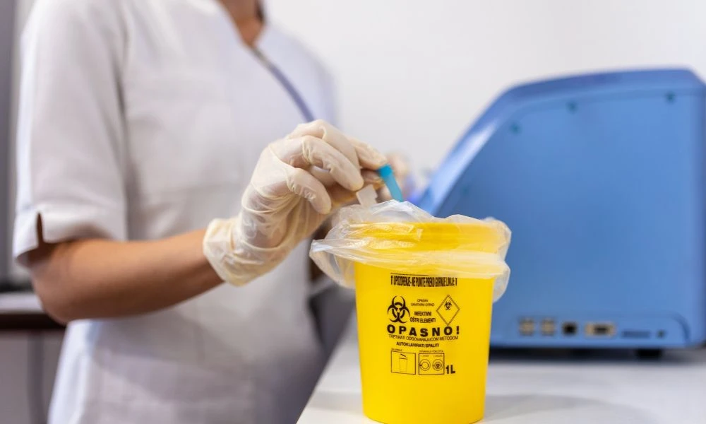 Professionnelle de soin portant des gants, manipulant une aiguille et une seringue au-dessus d'un conteneur jaune pour déchets à risques infectieux. Le conteneur porte l'inscription « DASRI ».