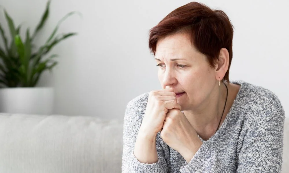 Femme assise sur un canapé, avec une expression inquiète ou troublée. Ses mains sont portées à sa bouche, une posture qui suggère de l'anxiété.