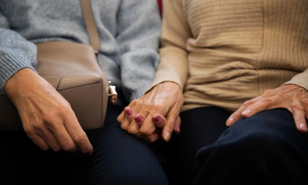 Deux dames âgées sont assises et se tiennent la main. L'une portent un sac à main et l'autre pas. On ne voit pas leur visage.