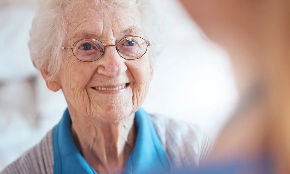 Dame âgée souriante portant des lunettes avec des montures fines aux cheveux blancs coiffés soigneusement. Elle fait face à une femme qui est en flou au premier plan.