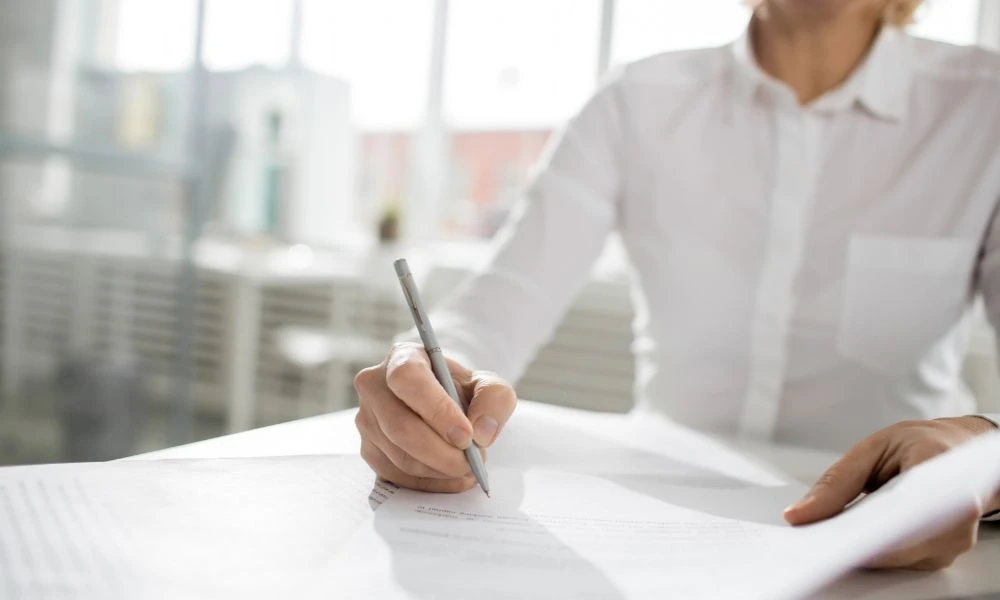 Une personne en chemise blanche signe un document posé sur une table lumineuse.