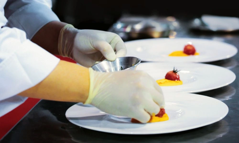 Mains gantées d'un cuisinier dressant des assiettes blanches garnies d'une tomate cerise sur une purée orangée, en cuisine professionnelle.