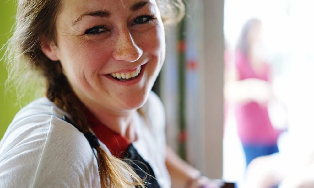 Une femme souriante regarde la caméra dans un environnement lumineux et convivial.