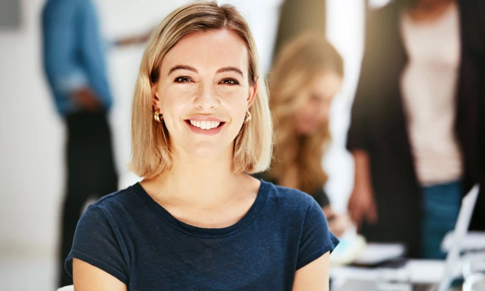 Une jeune femme souriante, assise dans un environnement de travail lumineux, regarde vers l’objectif.