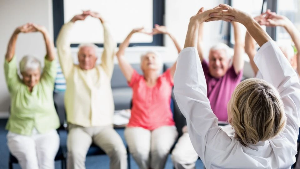 Groupe de personnes âgées assises sur des chaises, participant à une session d'exercices physiques. Elles sont dirigées par une personne debout, un professionnel de santé, située au premier plan et de dos.