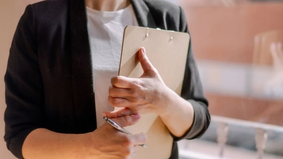 Une personne en veste noire et t-shirt blanc tient contre elle un porte-documents, prête à prendre des notes lors d'une inspection.