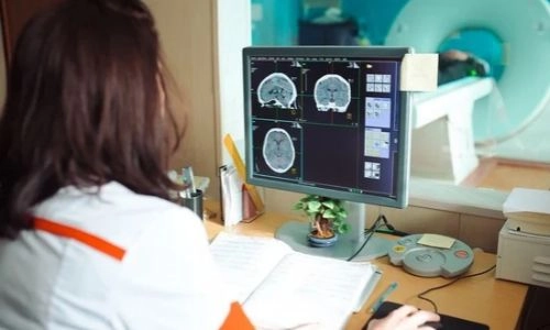 ne manipulatrice en radiologie analyse des coupes cérébrales de scanner sur un écran depuis la salle de commande, avec l'appareil d'imagerie visible derrière la vitre.