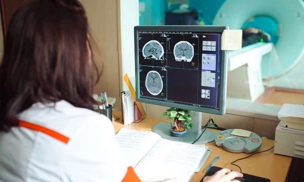 ne manipulatrice en radiologie analyse des coupes cérébrales de scanner sur un écran depuis la salle de commande, avec l'appareil d'imagerie visible derrière la vitre.