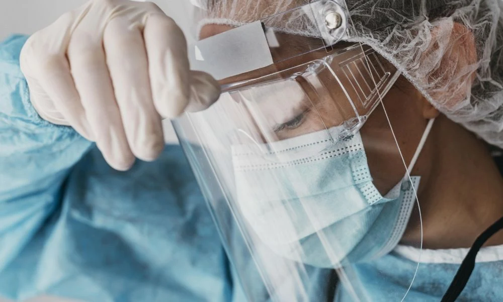 Un professionnel de santé en tenue de protection ajuste sa visière tout en portant un masque et des gants.